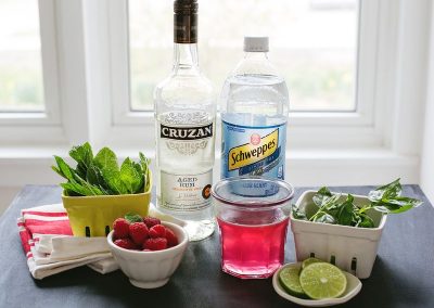 A bottle of vodka, lime, mint, and raspberries on a table.