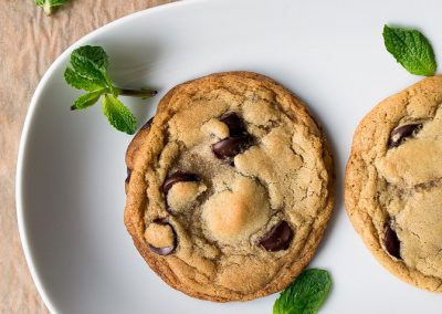 Two chocolate chip cookies on a plate decorated with mint leaves.